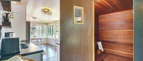 Una sala de sauna con paredes de madera y un banco, una zona de cocina con una cafetera y vistas al exterior a través de amplios ventanales.