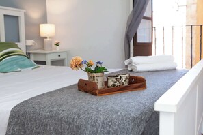 Un lit bien fait, recouvert d’une couverture grise, une table en bois ornée d’objets décoratifs, une lampe de chevet et une fenêtre avec des rideaux.