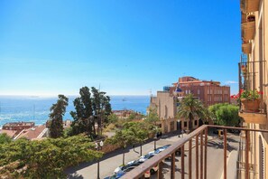 A coastal cityscape with a clear view of the sea, a street with parked cars, and buildings with balconies.