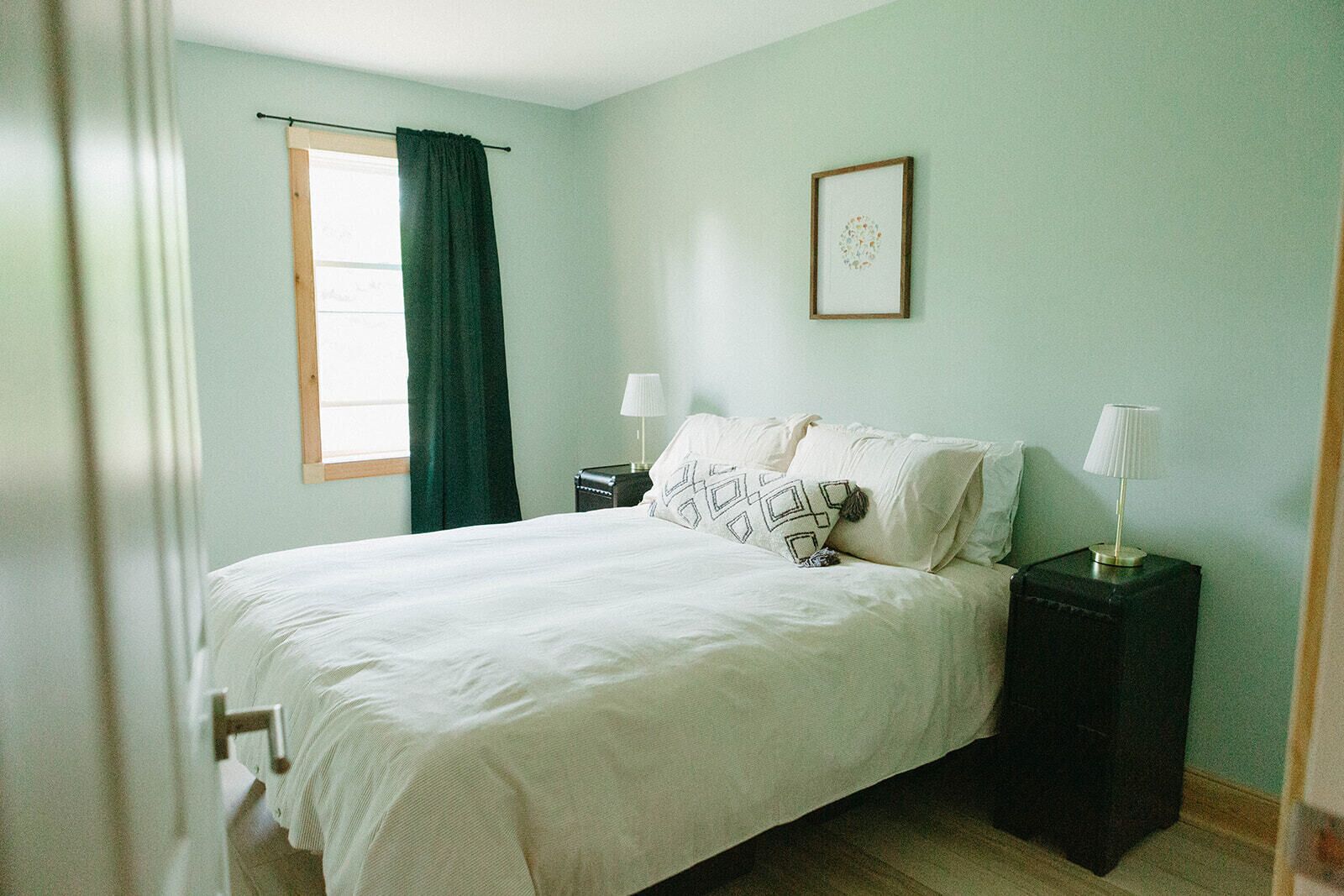 A bedroom with a bed, two bedside tables, a window with curtains, and a framed picture on the wall.