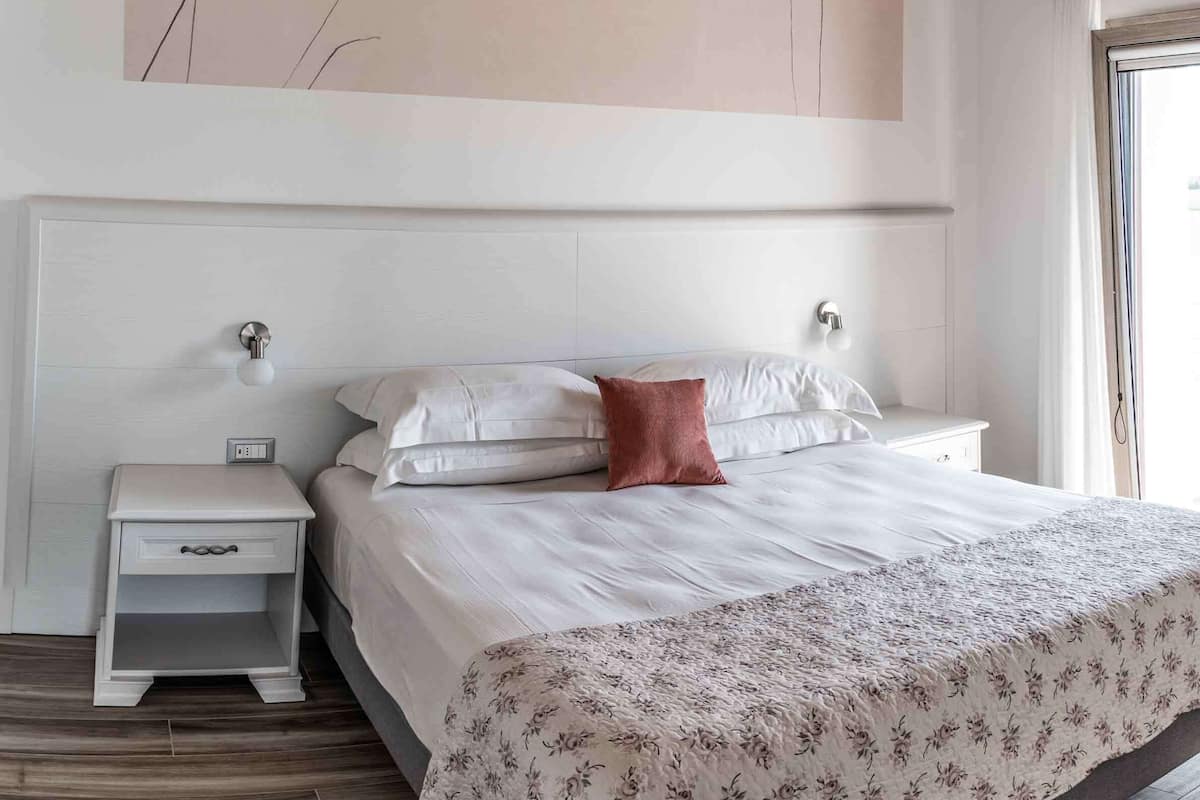 Um quarto com uma grande pintura abstrata, uma cama com roupa de cama branca e colcha florida, uma mesa de cabeceira com gaveta e uma janela com cortinas.