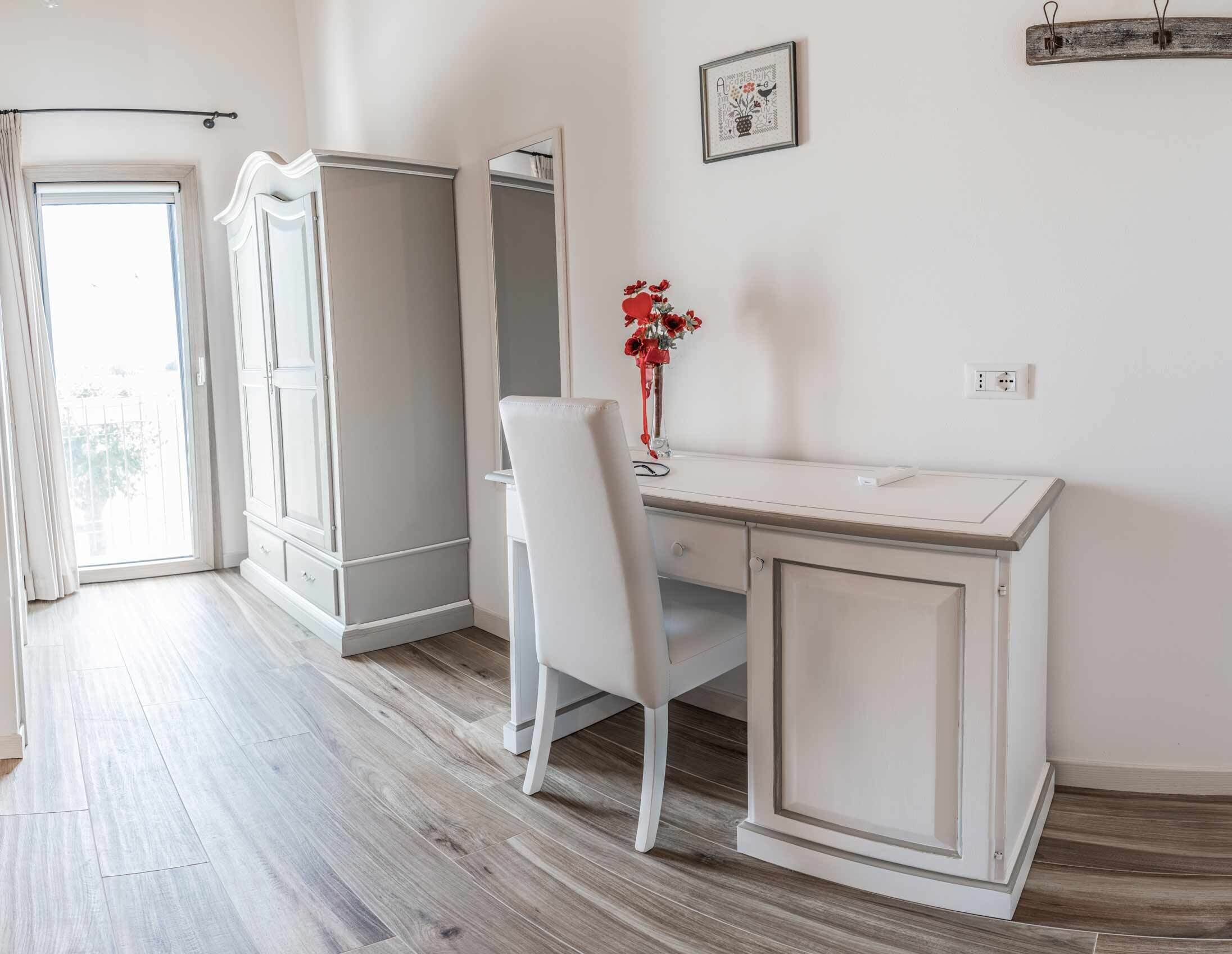 A room with a white desk, chair, and wardrobe, a small red vase with flowers, and a framed picture on the wall.