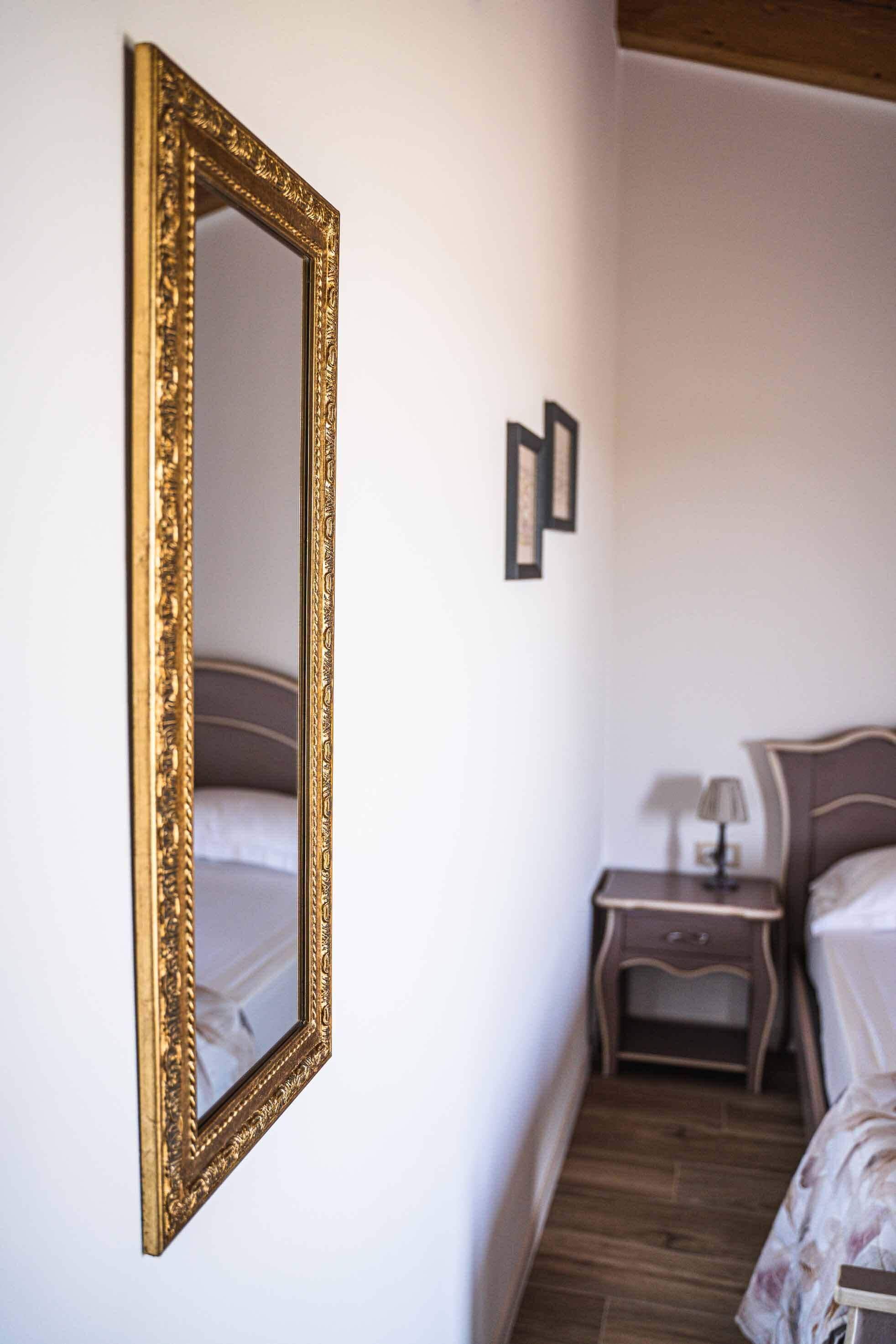 A bedroom with a large ornate mirror, a bed with a floral bedspread, a nightstand with a lamp, and a wooden headboard.