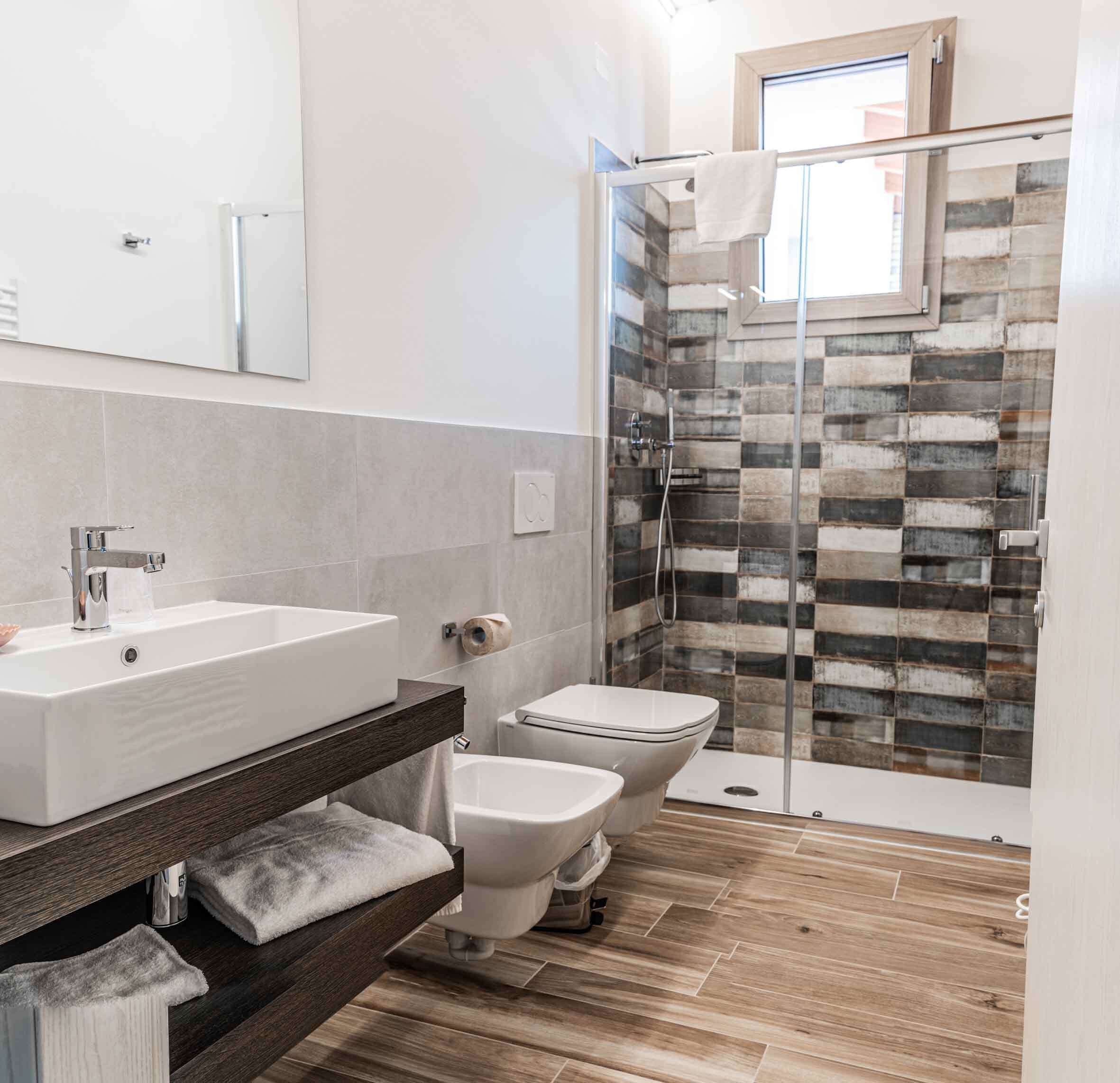 A modern bathroom with a large white sink, two toilets, a glass shower enclosure, and a wooden floor.