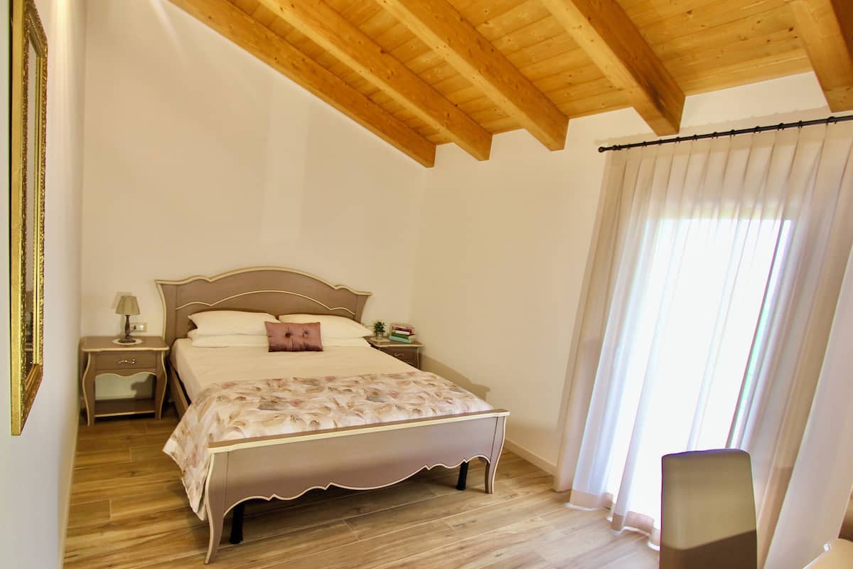 Quarto com teto de madeira, cama com colcha floral, mesa de cabeceira com abajur e janela com cortinas.