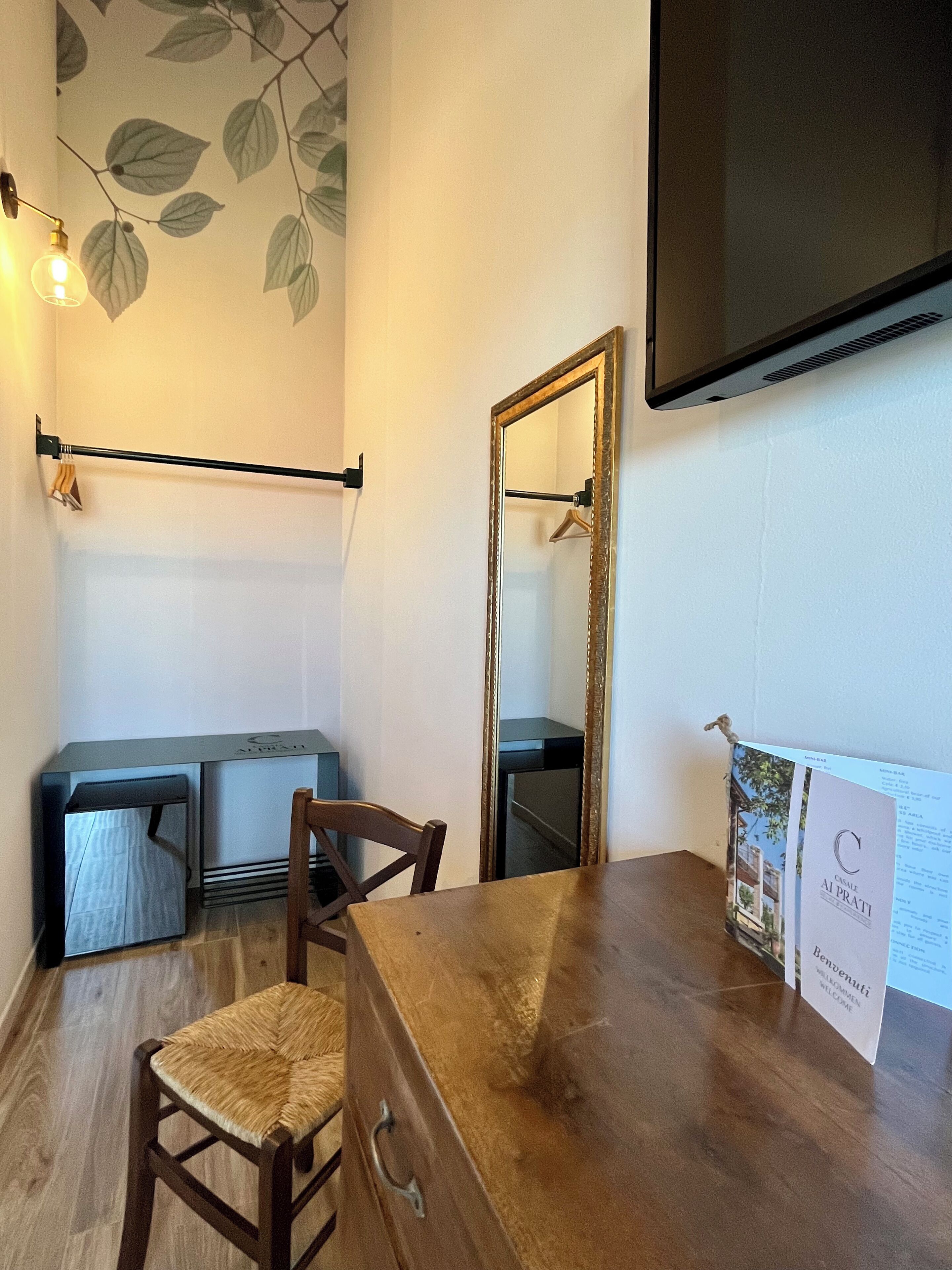 A room with a wooden desk, a chair, a hanging coat rack, a mirror, and a wall-mounted television.
