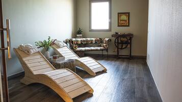 Een kamer met twee houten lounge fauteuils, een bank met bloemenprint, een kleine tafel met een plant en een raam dat natuurlijk licht binnenlaat.