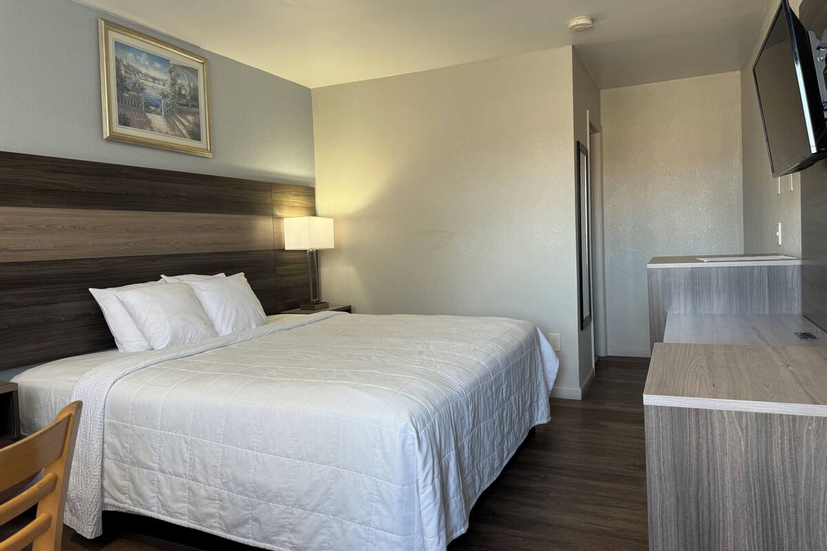 A hotel room with a large bed, a bedside table with a lamp, a wooden headboard, a painting on the wall, and a TV mounted on the wall.