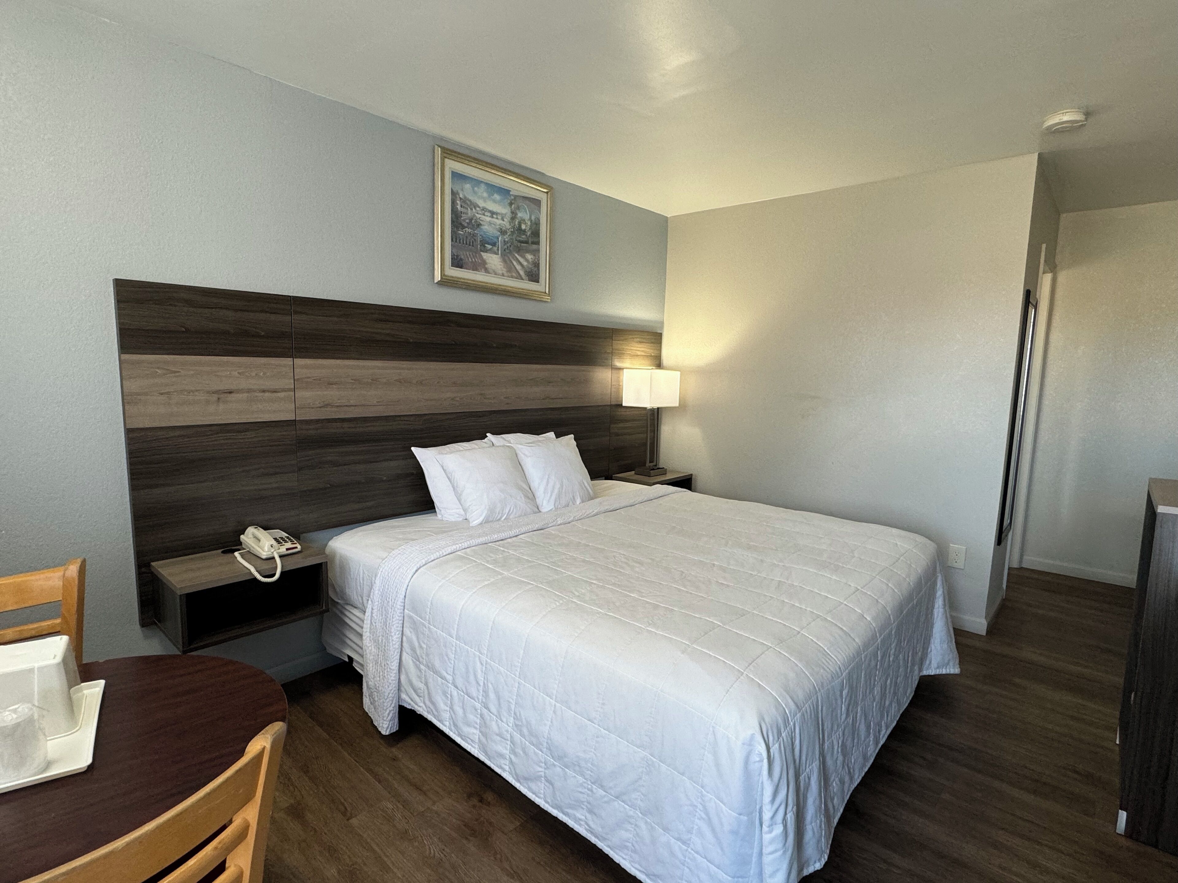 A hotel room with a large bed, a wooden headboard, a bedside table with a lamp, a phone, and a painting on the wall.