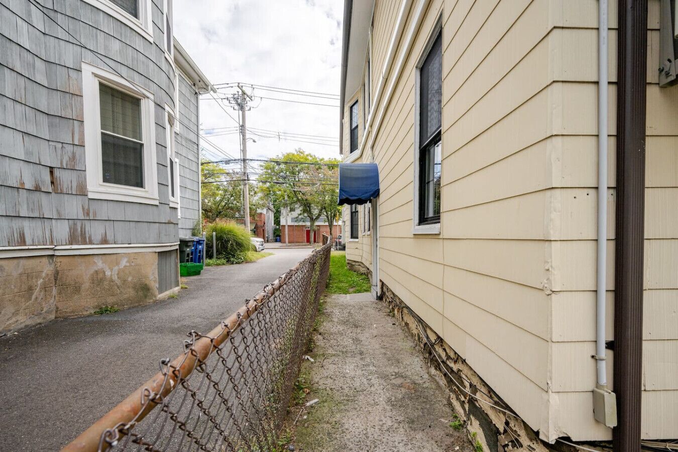A narrow alleyway between two houses with a chain-link fence and a blue awning on one side.