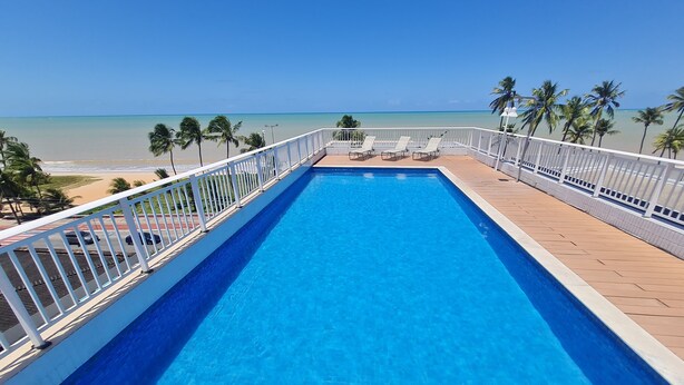 A rectangular pool with a white border, surrounded by a wooden deck and white railings. - JARDINS DO ATLANTICO by Penareia.tur.br (João Pessoa)