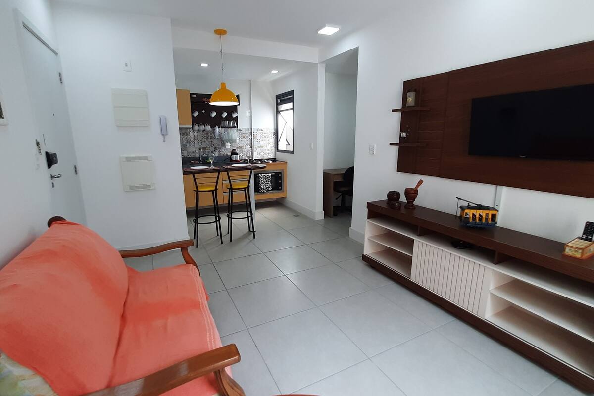 Un salon moderne avec un canapé, un téléviseur fixé au mur et une cuisine en arrière-plan.