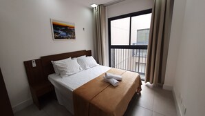 Una cama bien hecha con ropa blanca y una manta marrón, un cuadro pequeño en la pared y una ventana con cortinas.