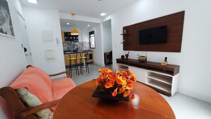 Una sala de estar moderna con sofá, una mesa de comedor con un florero, una zona de cocina con barra y un televisor montado.