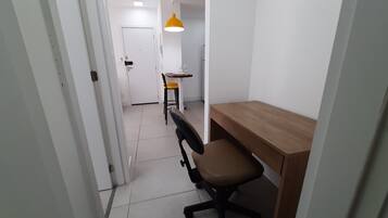 A narrow hallway with a wooden desk and office chair, a yellow pendant light, and a white door at the end.