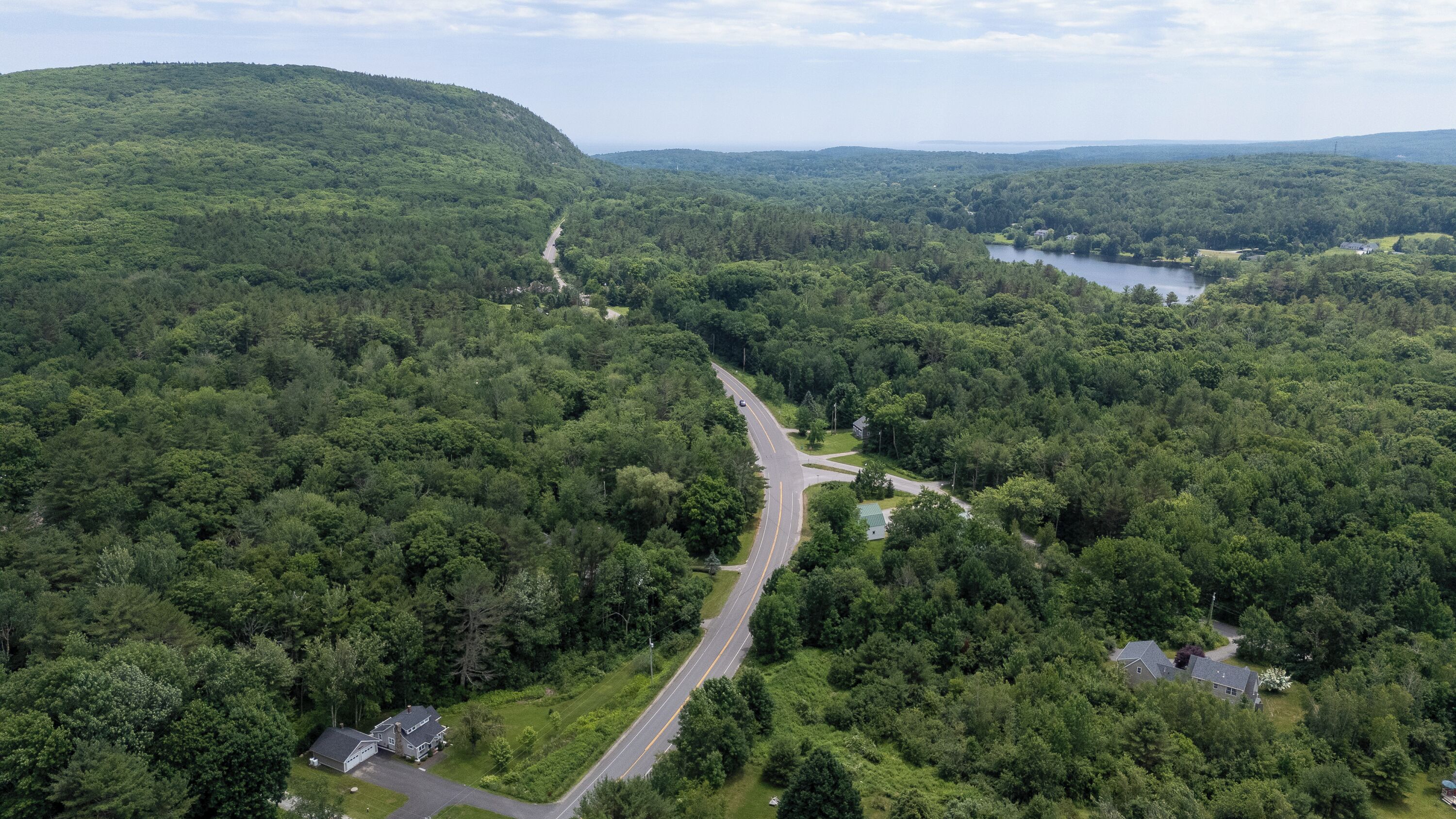 An aerial view of a forested area with a winding road, a lake, and a hill.