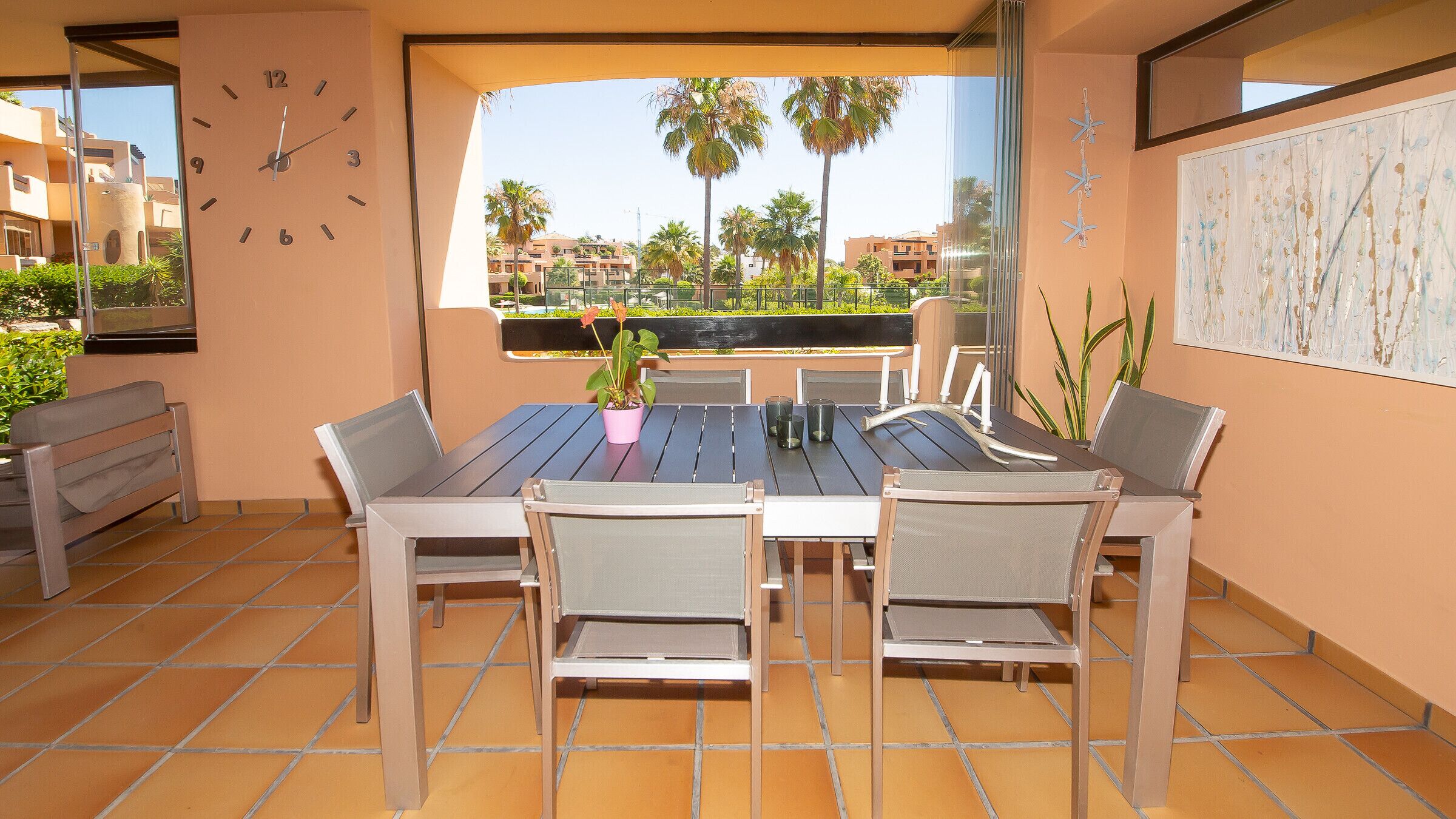 Un patio con mesa y sillas, un reloj y vistas a palmeras y edificios.