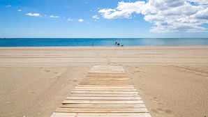 Ein Holzsteg führt zu einem Strand mit klarem, blauem Wasser und einigen Personen in der Ferne.