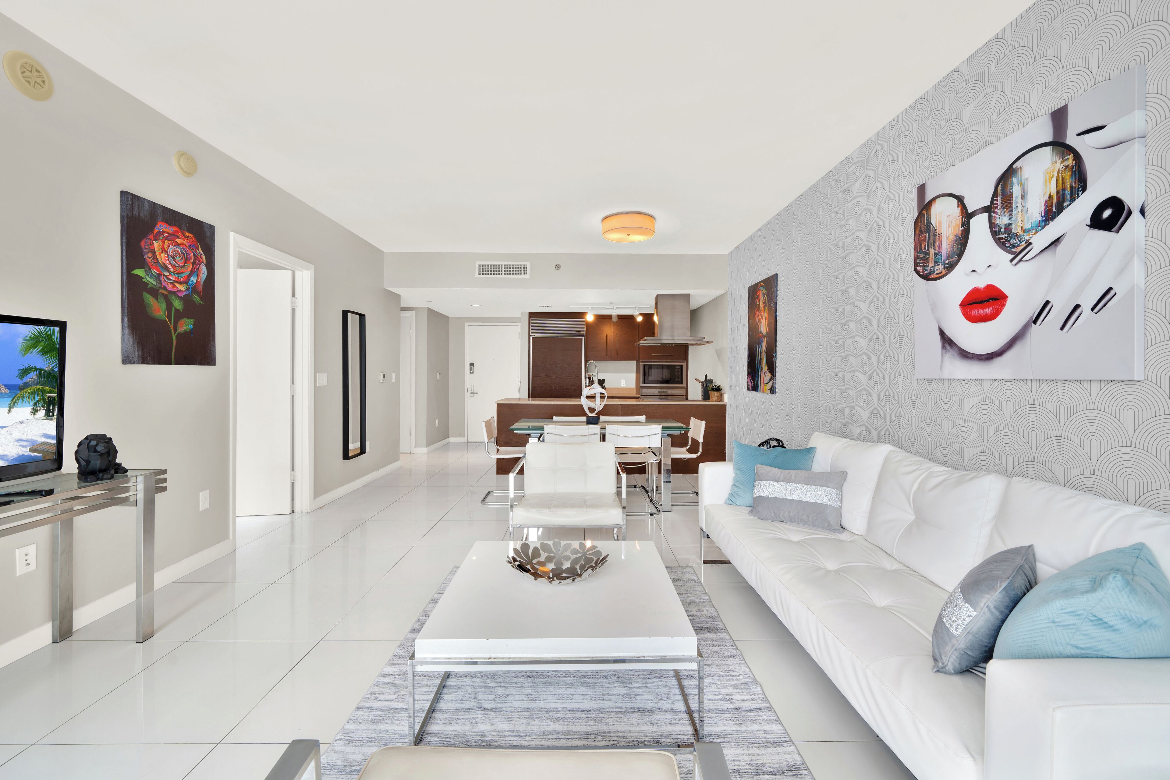 A modern living room with a white sofa, a coffee table, and a TV. There is a dining area with a table and chairs, and a kitchen in the background.