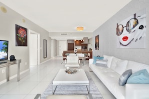 A modern living room with a white sofa, a coffee table, and a TV. There is a dining area with a table and chairs, and a kitchen in the background.