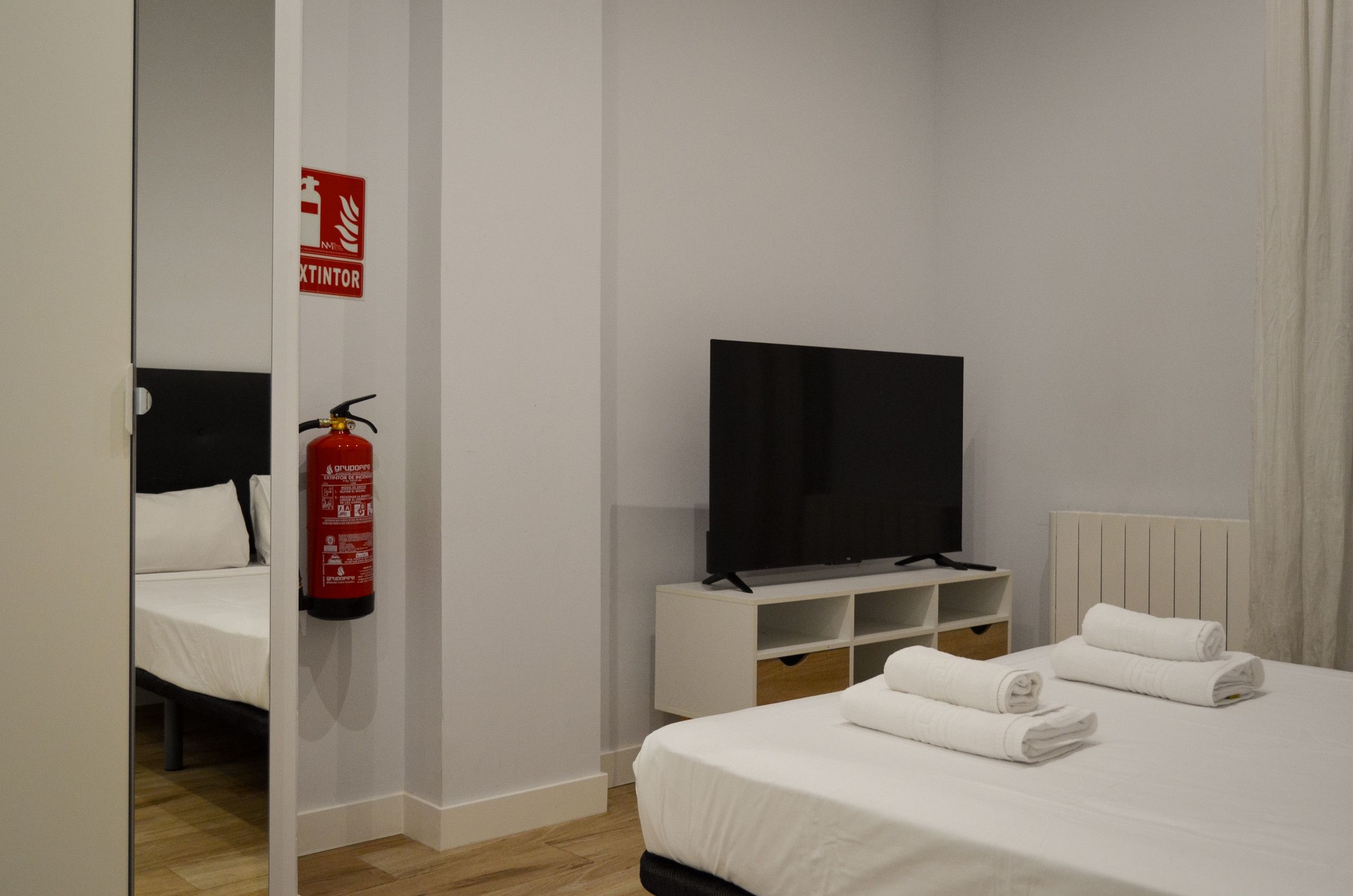 Een hotelkamer met een bed, een televisie op een standaard, een brandblusser en een spiegel.