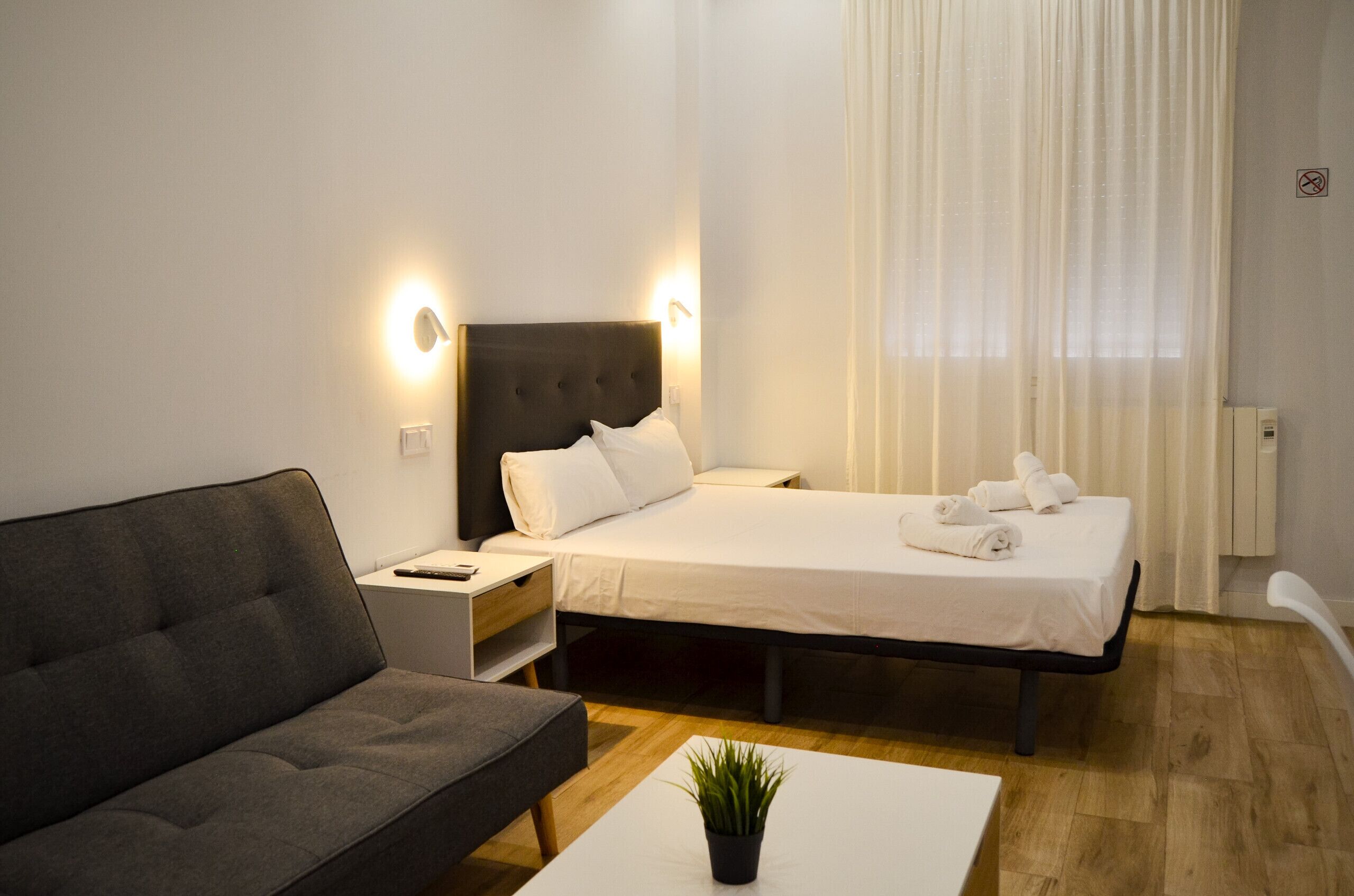 Una habitación de hotel con una cama, una mesita de noche, un sofá, una pequeña mesa con una planta y una ventana con cortinas.