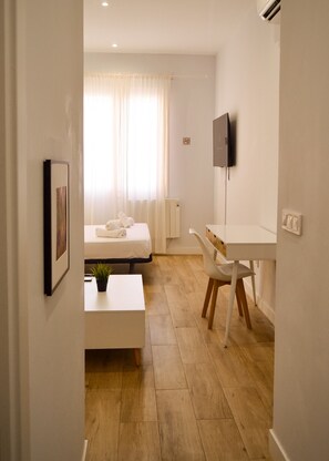 A hotel room with a bed, a desk, a chair, a small table with a plant, and a television on the wall.
