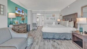 A hotel room with a large bed, a desk, a chair, and a dresser. There are wall art and a lamp on the dresser. - The Caravelle Resort (Myrtle Beach)