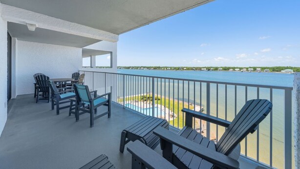 A balcony with Adirondack chairs, a table, and a view of a body of water with a distant shoreline.