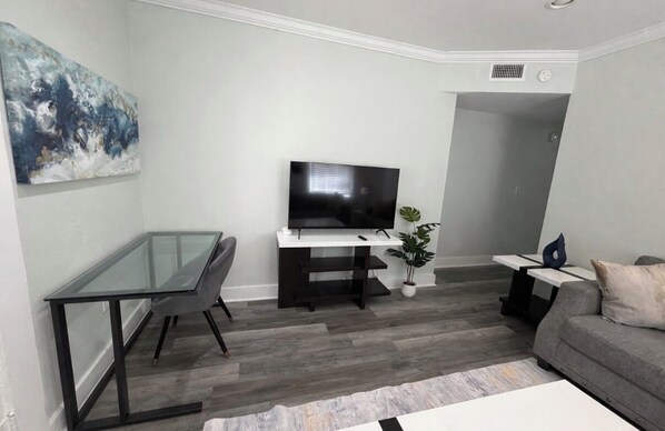 A modern living room with a glass-top table, a black sofa, a flat-screen TV, and a large abstract painting on the wall. - Thornton park oasis 1 bedroom (Orlando)