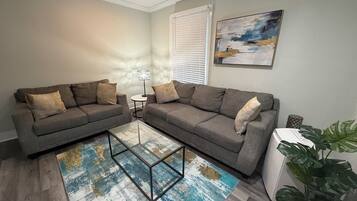 A living room with two grey sofas, a glass coffee table, a patterned rug, a painting on the wall, and a potted plant.