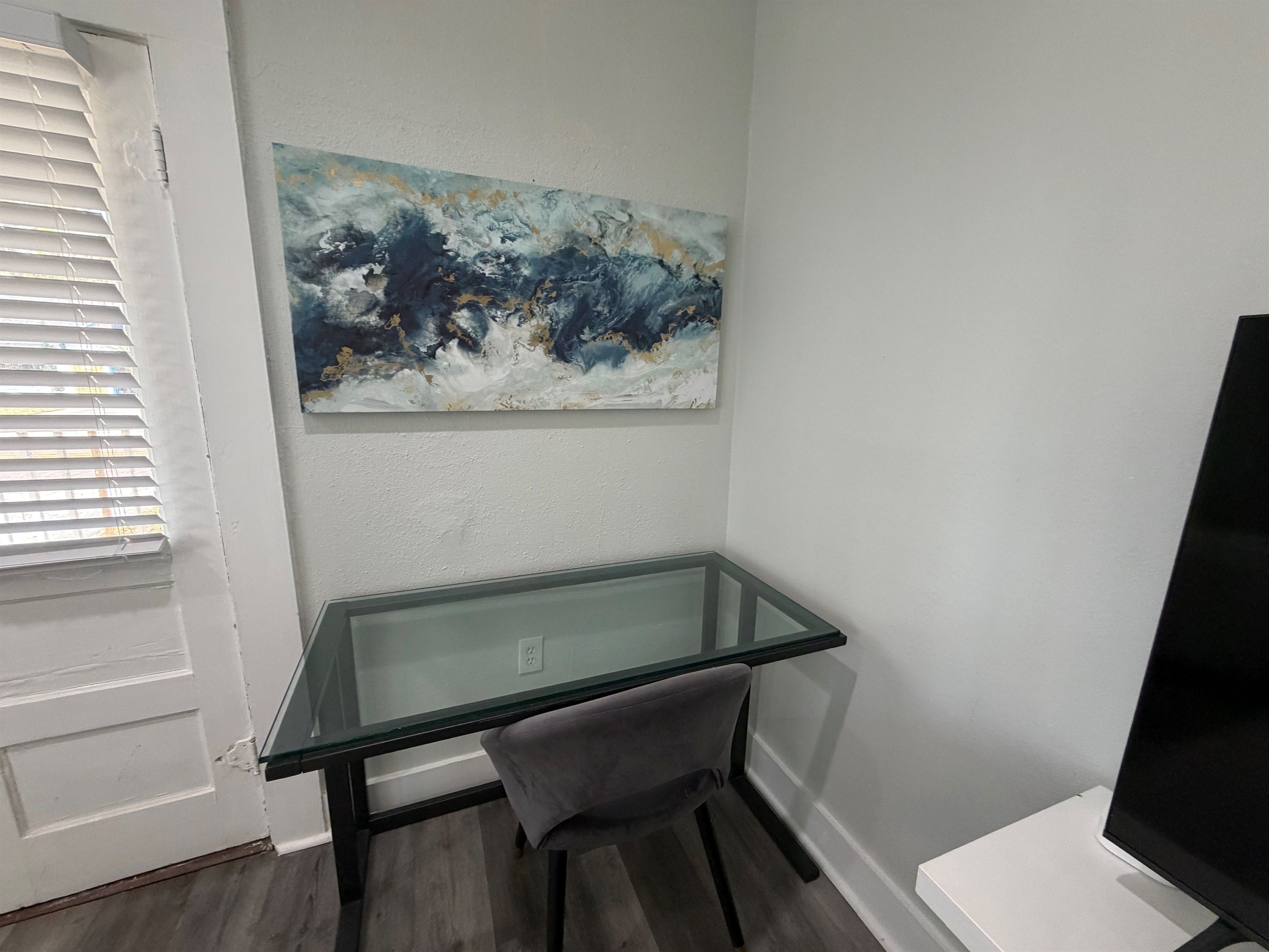 Une pièce avec un bureau en verre, une chaise, un tableau accroché au mur et un téléviseur posé sur un meuble.