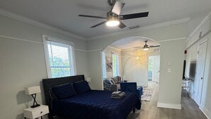 A bedroom with a bed, a desk, and a chair. There is a ceiling fan and a window with blinds.