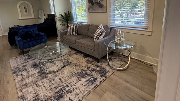 A living room with a grey sofa, a blue armchair, a glass coffee table, a patterned rug, and a chandelier.