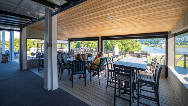 An outdoor dining area with wooden tables and chairs, large windows, and a view of a lake.