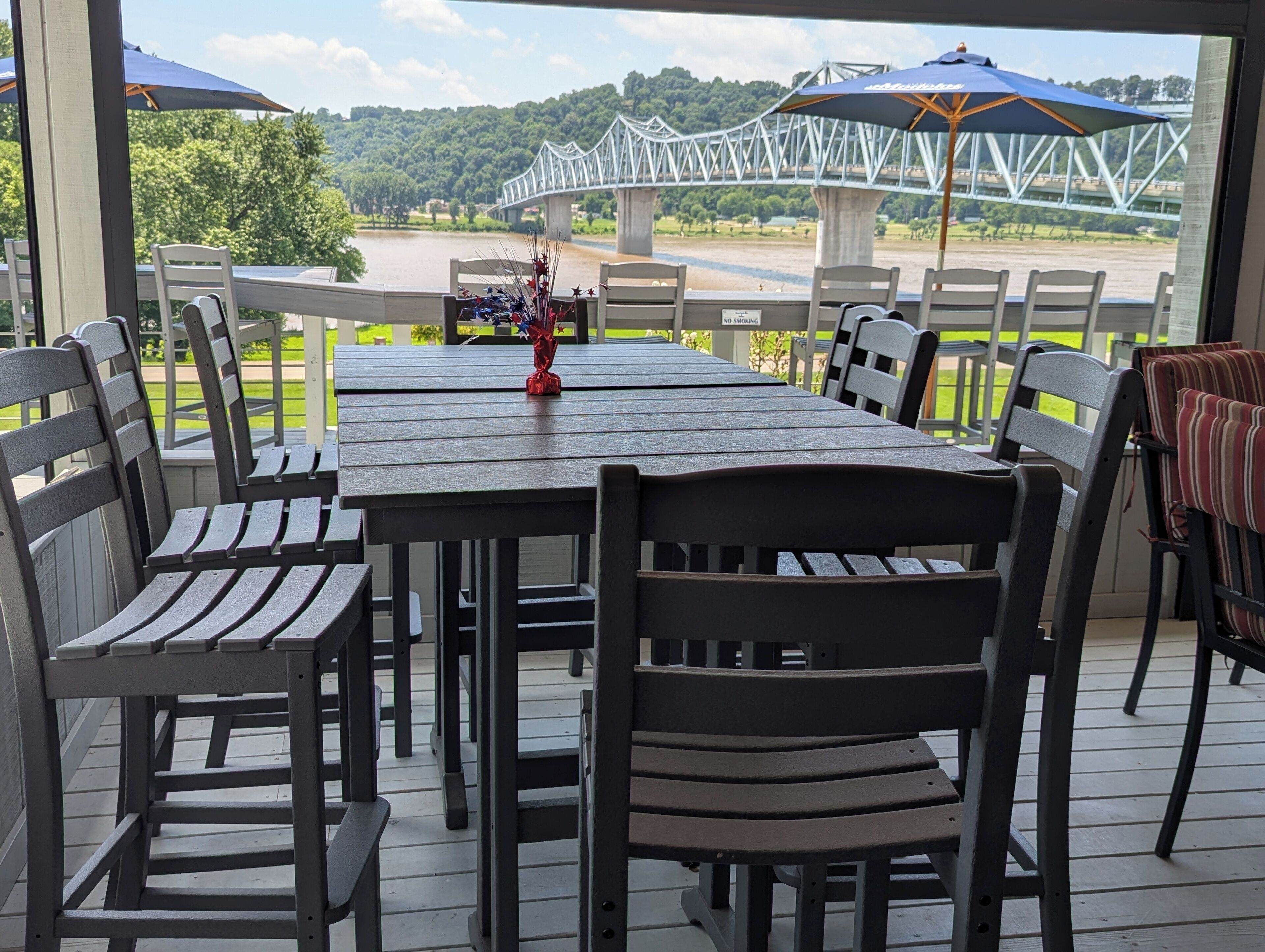 An outdoor patio with a long table and chairs, a decorative vase, and a view of a bridge and river.