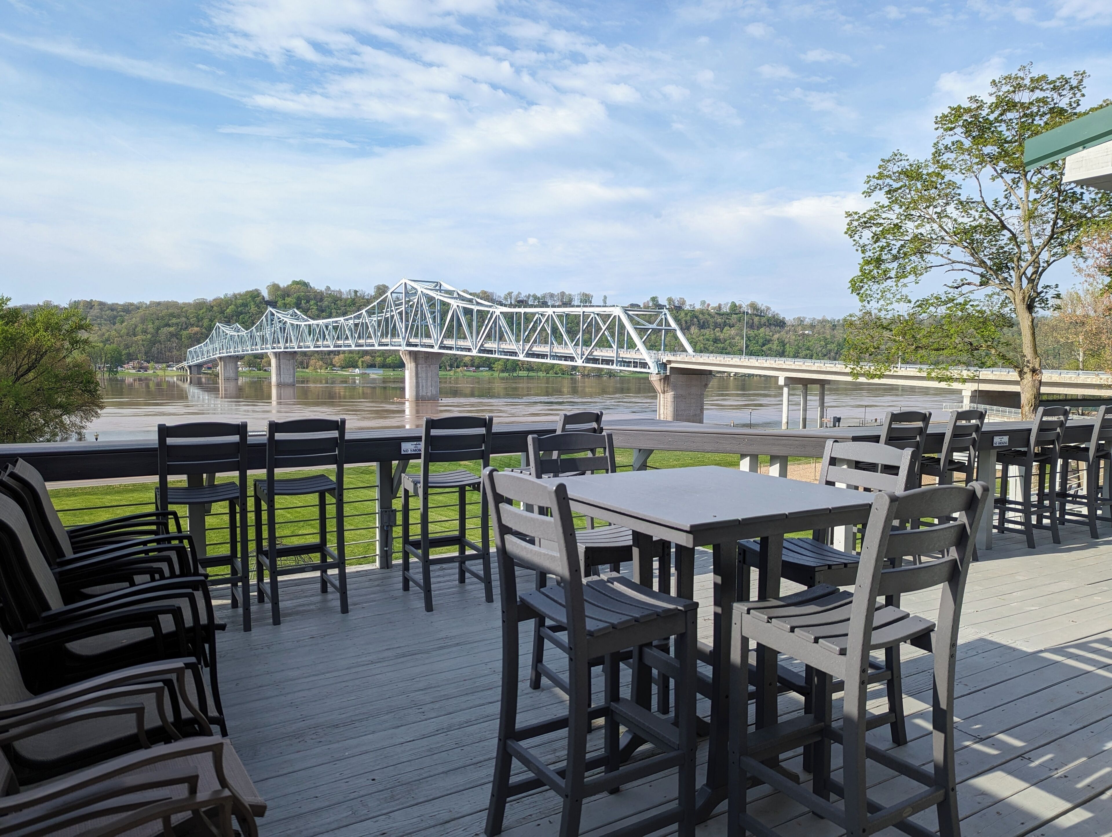 An outdoor seating area with tables and chairs, overlooking a river and a bridge.