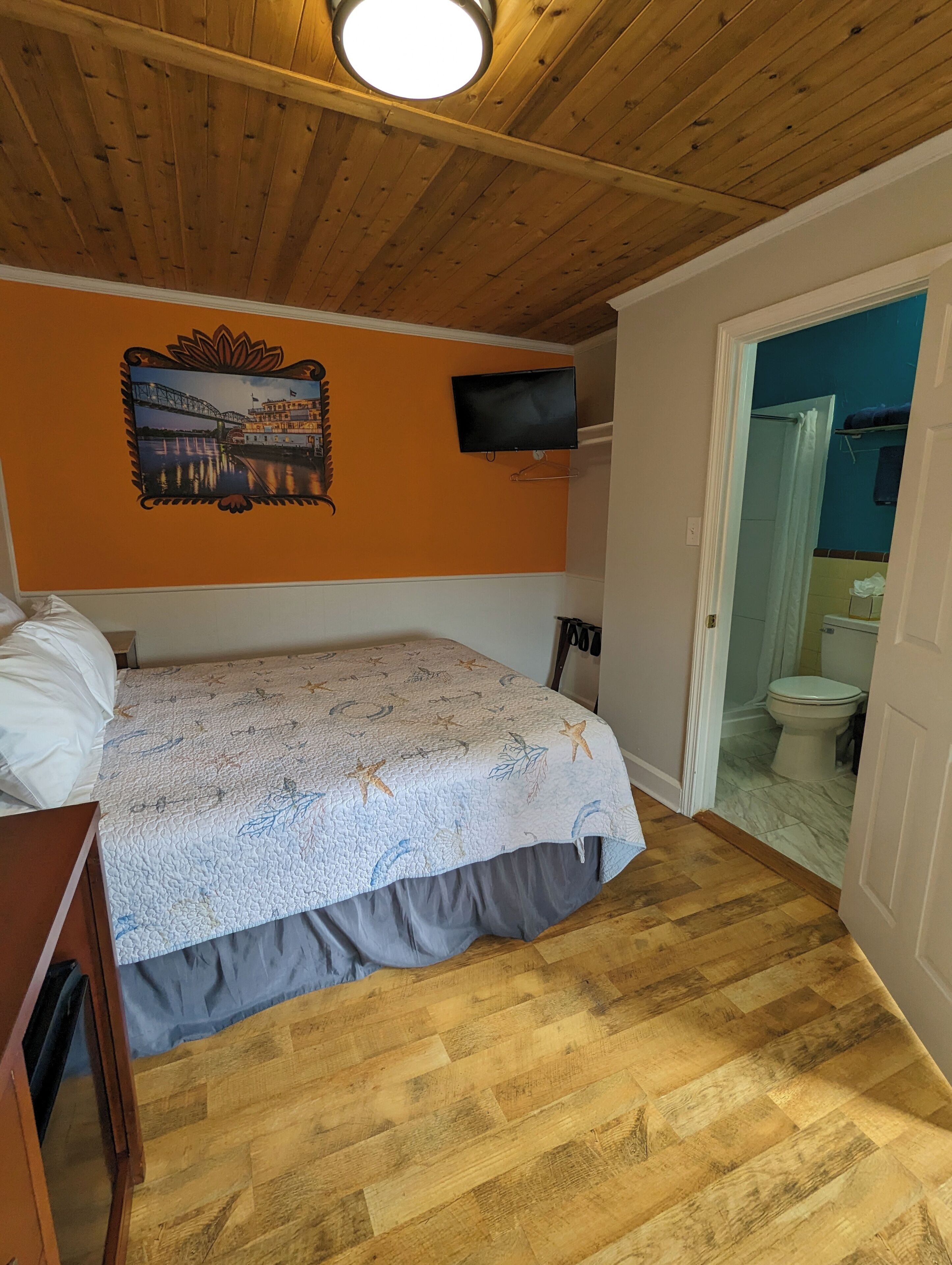 A room with a bed, a mounted TV, and a wooden ceiling.