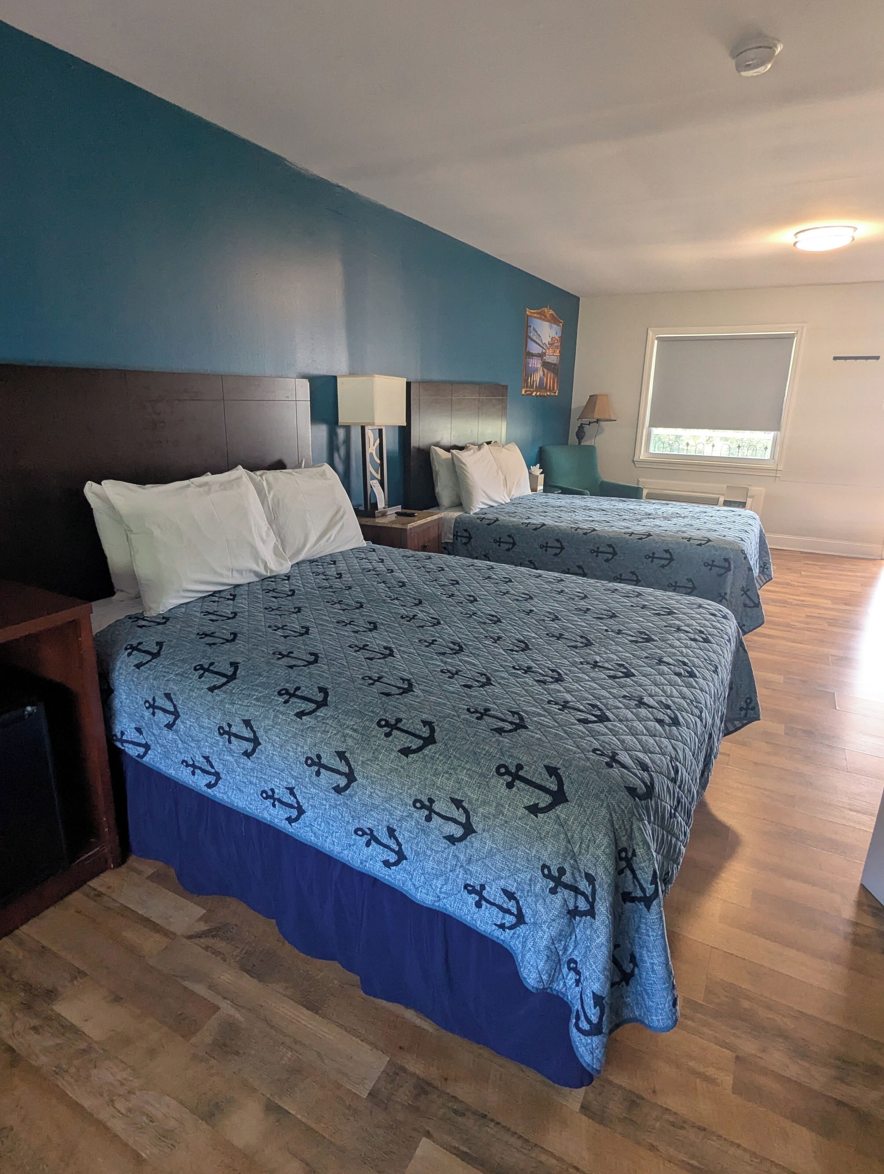 A hotel room with two beds, wooden flooring, a nightstand, a lamp, and a window with a view.