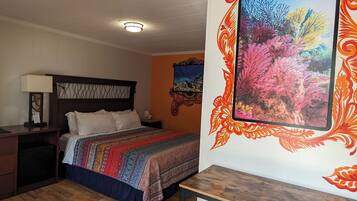 Una camera d'albergo con un letto, una scrivania con degli sgabelli, un comodino, una lampada e un quadro colorato appeso al muro.
