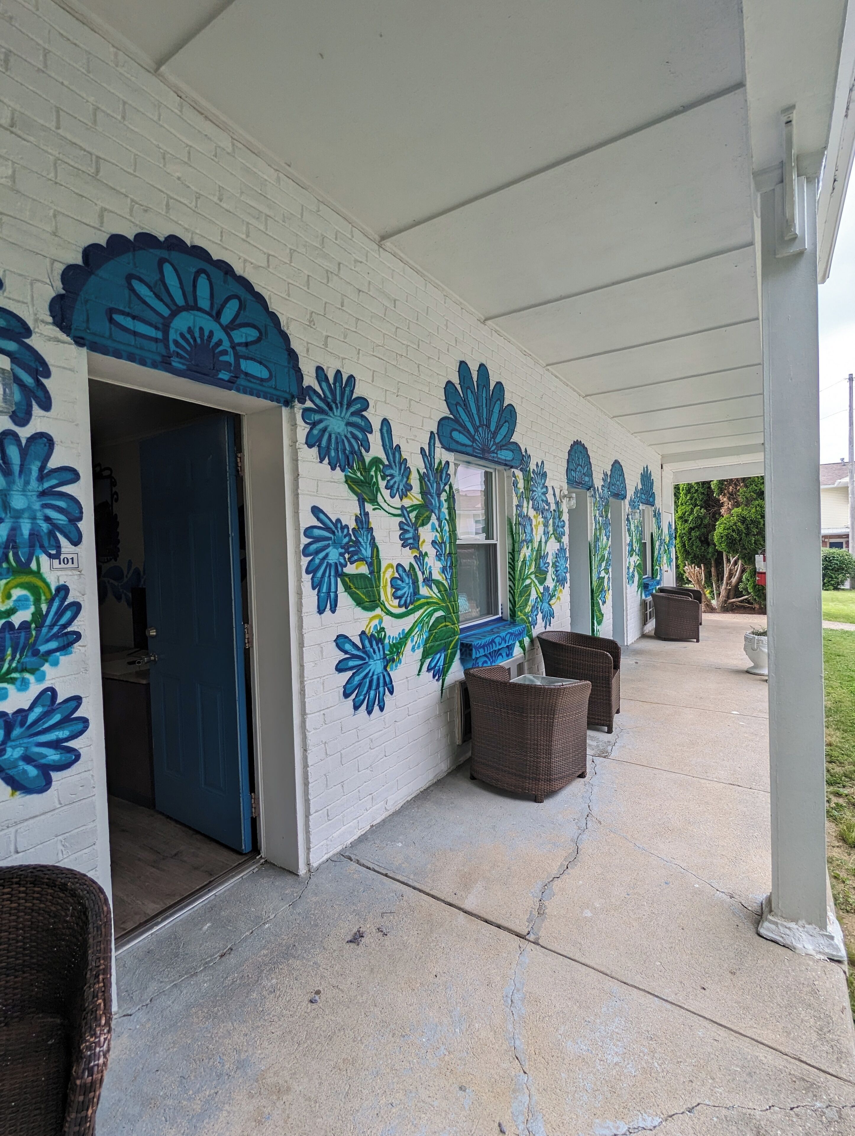 A covered outdoor area with a white brick wall featuring blue floral murals, wicker furniture, and a concrete floor.