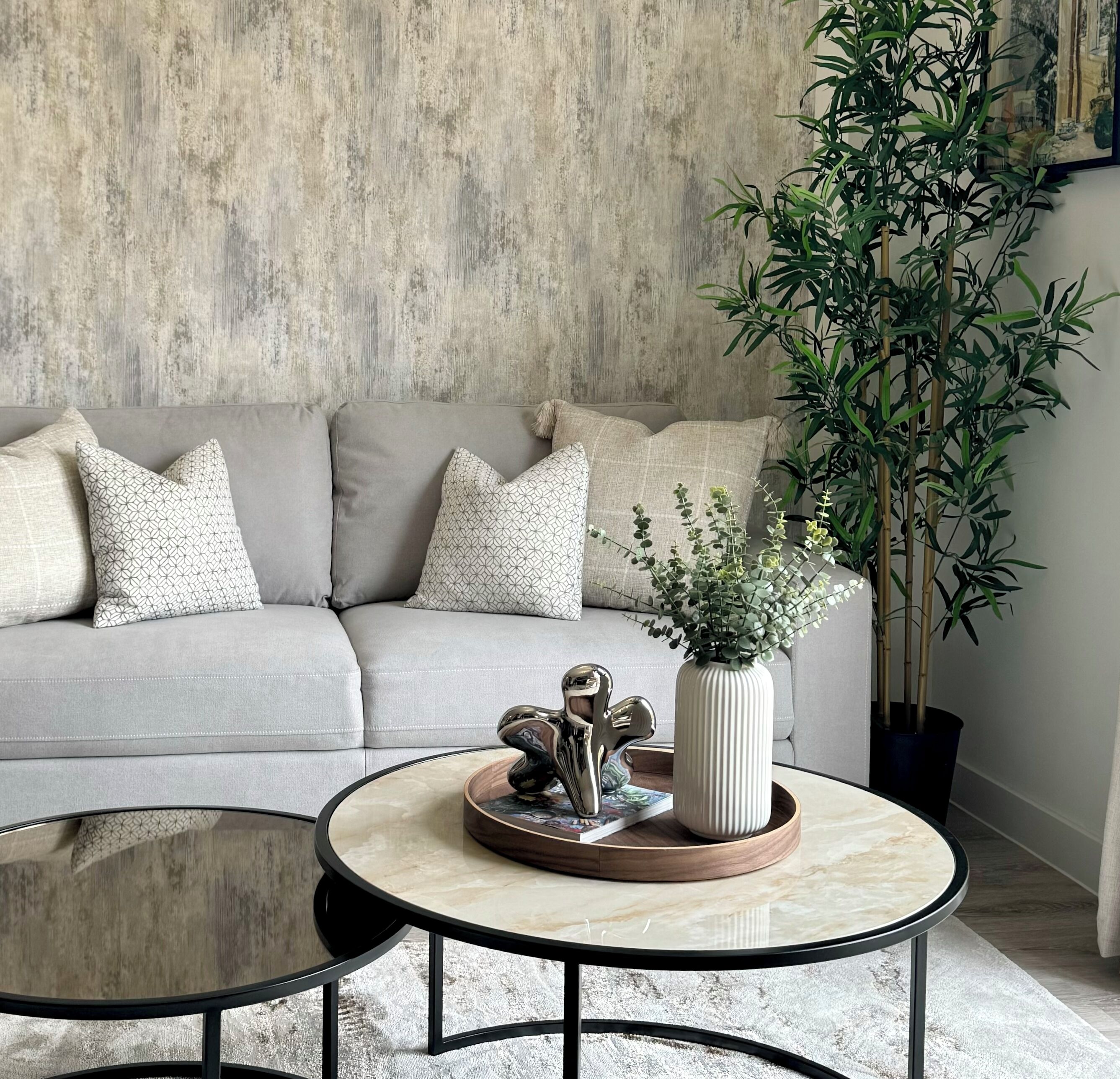 Un salón con sofá gris, cojines estampados, una mesa redonda de centro con una escultura metálica decorativa, un jarrón blanco con plantas verdes y una planta de bambú alta.