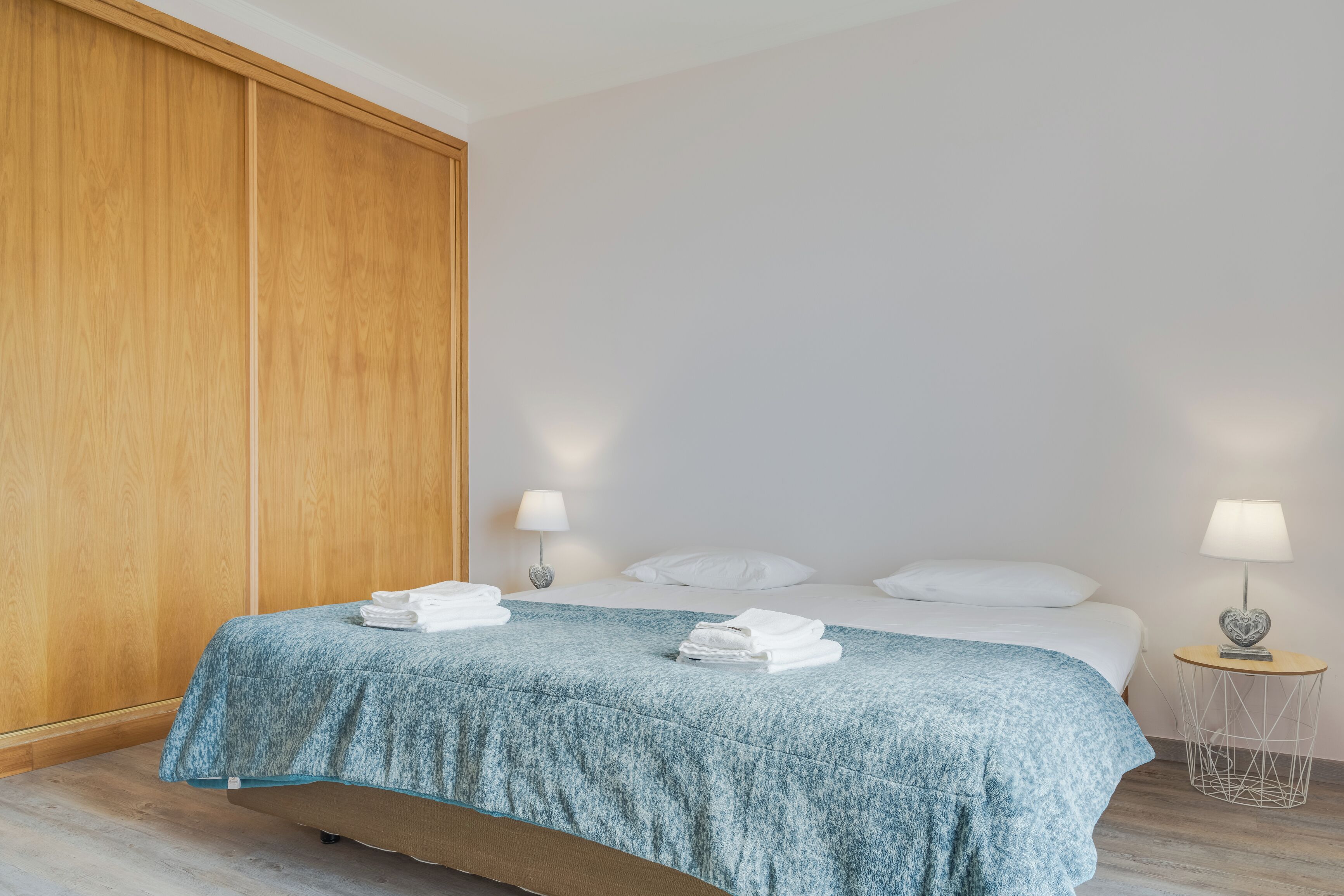 A neatly made bed with a blue bedspread, two bedside lamps, and a wooden wardrobe.