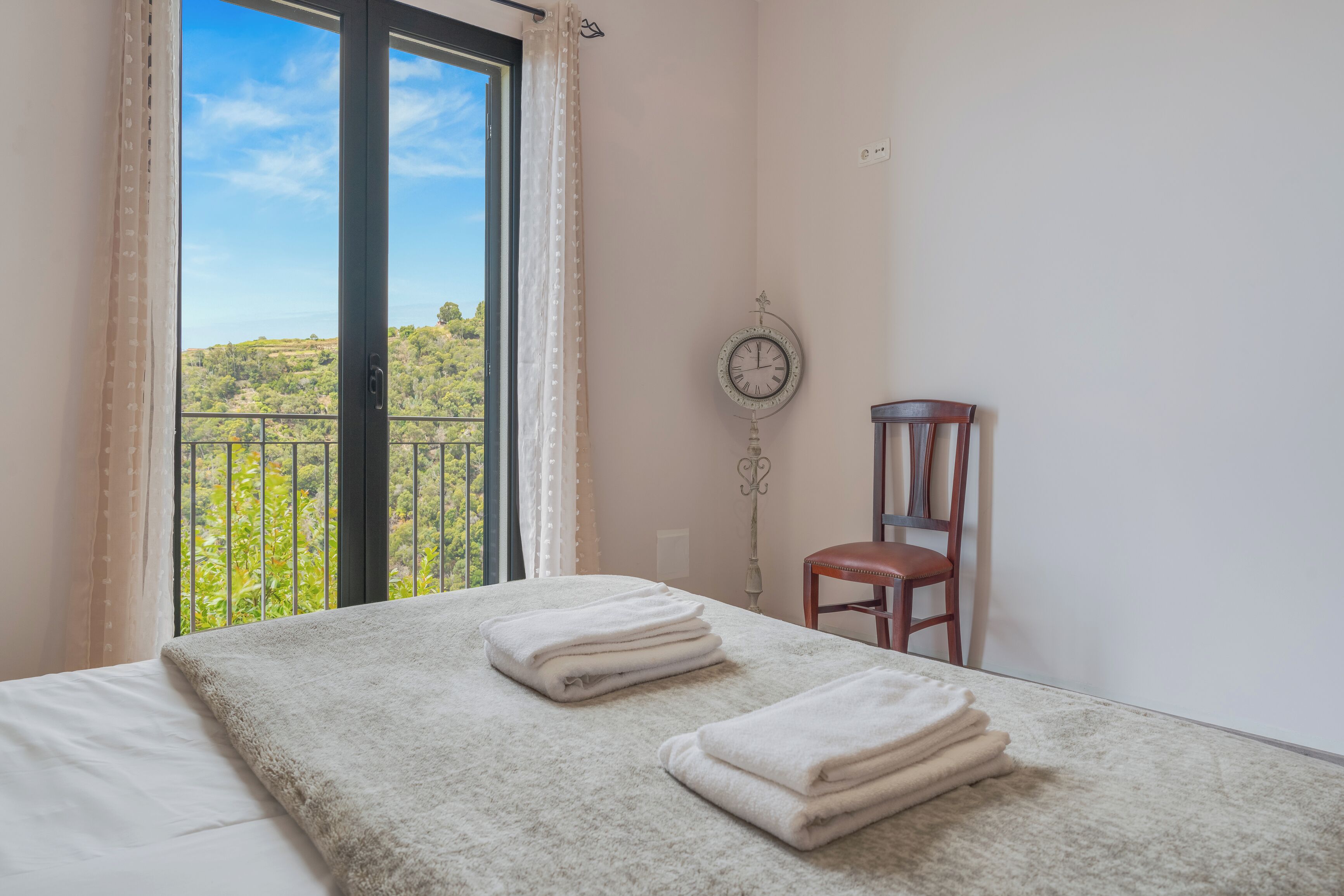A bedroom with a bed, a chair, a clock, and a view of greenery outside.