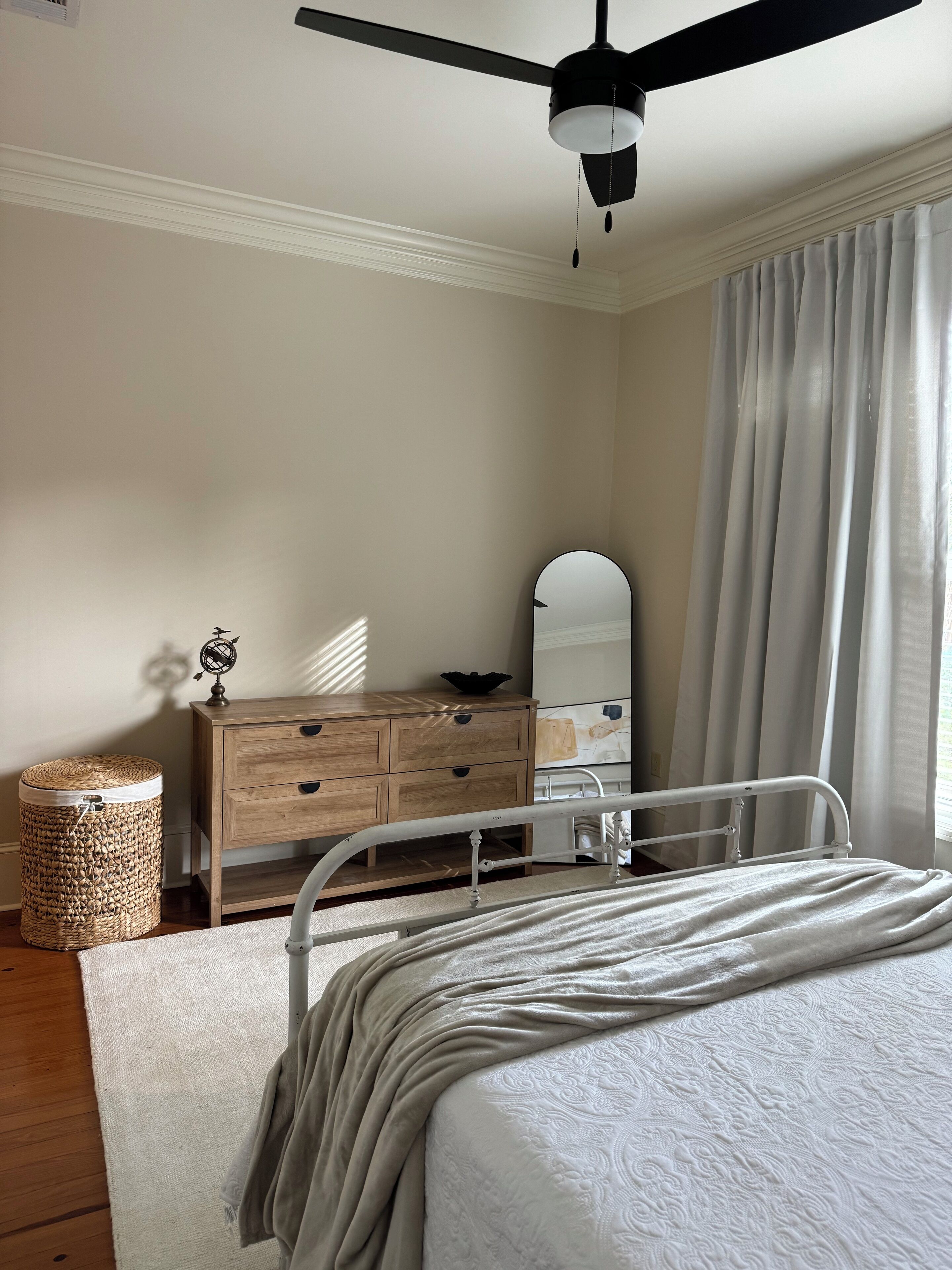 A bedroom with a bed, a wooden dresser, a mirror, a ceiling fan, and a window with curtains.