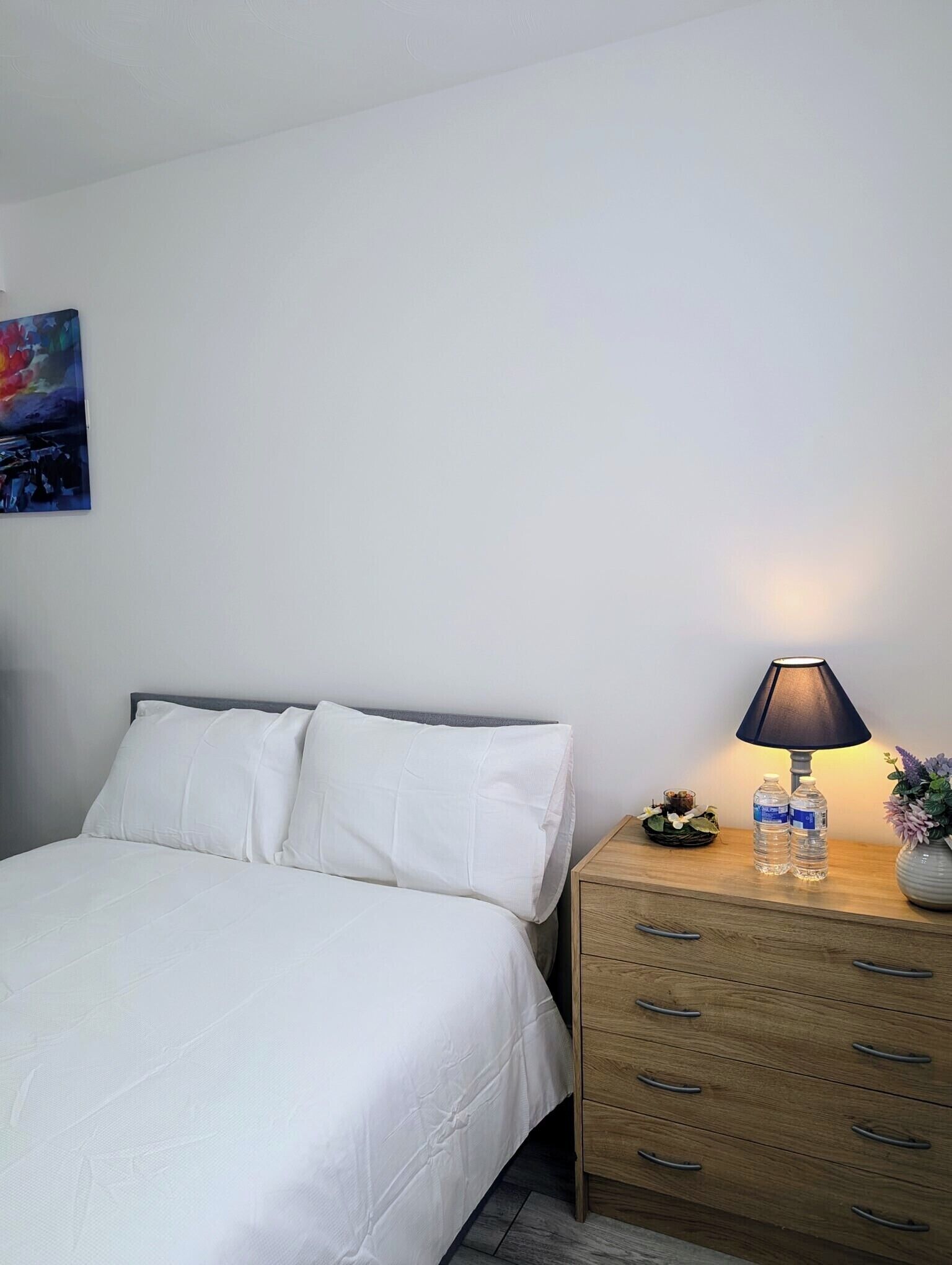 Un lit bien fait, avec du linge de lit blanc, une table de chevet en bois surmontée d’une lampe et une petite plante décorative.