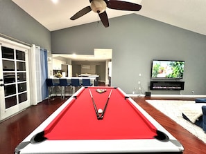 Mesa de bilhar numa sala com ventoinha no teto, televisão e zona de refeições ao fundo.