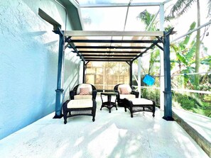 Uma área de estar coberta ao ar livre com móveis de vime e uma porta de vidro que dá para um jardim exuberante.