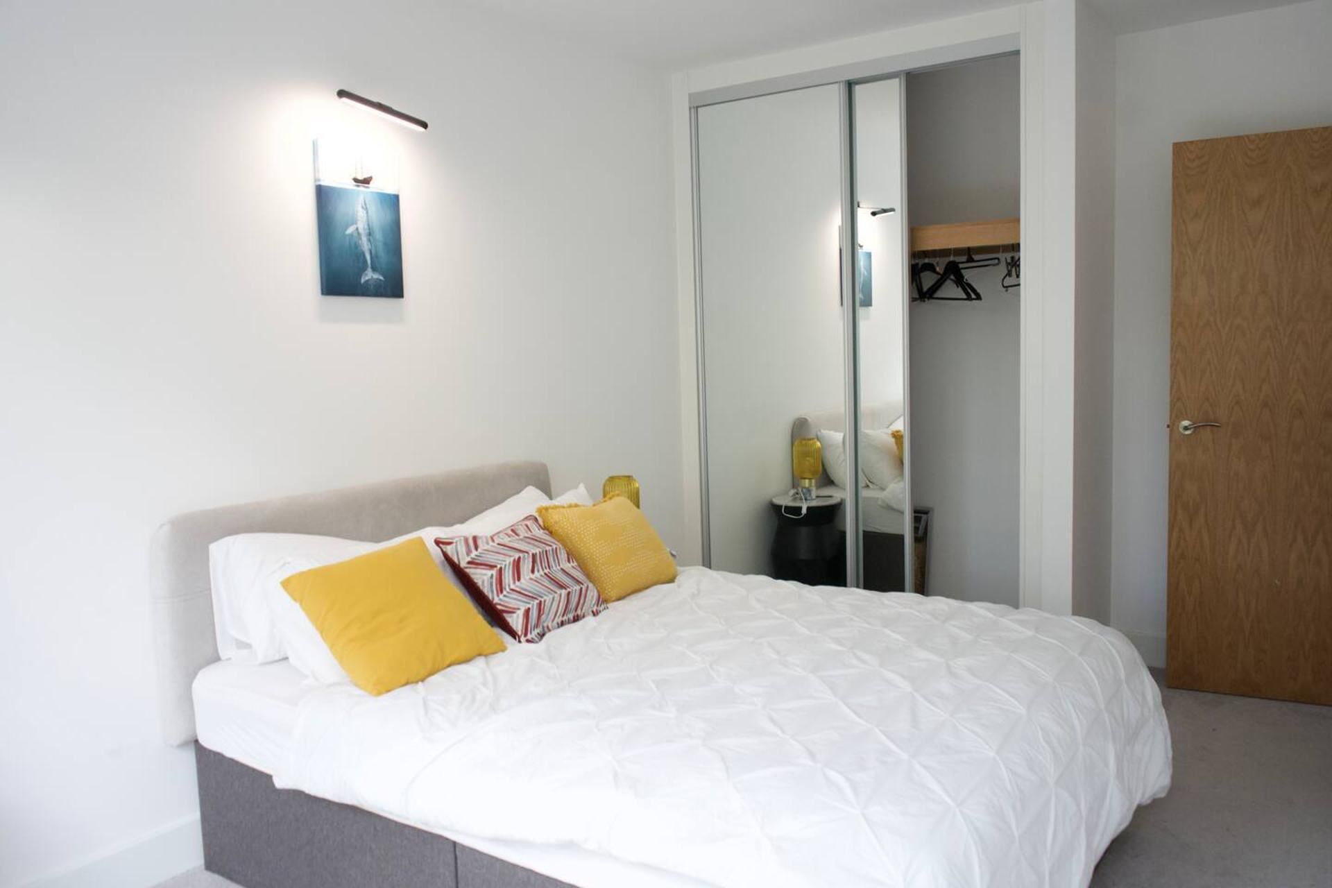A neatly made bed with white bedding and two pillows, one yellow and one with a red and white pattern. A small bedside table with a lamp is visible.