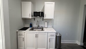 Una cocina compacta con muebles blancos, un lavamanos de acero inoxidable, un microondas y un cubo de basura.
